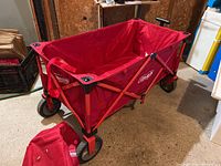 Red Coleman fold-up canvas wagon positioned in a garage area with surroundings including storage boxes and other household items.