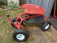 Full side view of red metal garden seat with four large treaded wheels on grass next to a shed.