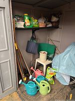 Photo showing gardening tools, watering cans in various colors, fertilizer, baskets, and a plastic stool inside garden shed.