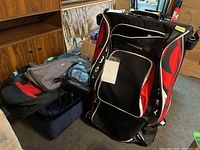 Front view of large black, red, and white Grit hockey tower bag showing main compartment and front pocket with visible Grit branding. Additional bags seen piled next to it: gray/red soft bag, black leather bag.