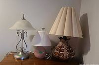 Three table lamps on wooden surface: a tall metal lamp with frosted glass shade, a small pink ceramic lamp with pleated white shade, and a pottery lamp with leaf designs and large pleated beige shade.
