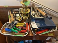Top view of multiple trays containing wire brushes, screwdrivers, pliers, tin snips, and measuring tape with a brass lamp in background.