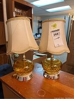 Pair of amber glass lamps with beige fabric shades on a wooden surface under bright lighting. Lamps have slender necks and circular metal bases.