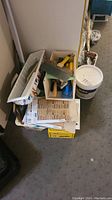 Photo showing stacked plastic containers and boxes with various painting tools, sandpaper, a saw, and a 5-gallon bucket of paint in a basement setting.