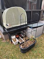 Wide shot showing large beige resin Vegepod container with mesh cover, black resin planters with trellis supports, ceramic pots in resin trays, and garden tools organized outside on grass near deck.