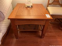 Front view of the wood side table with lamp on top, showing the pull-out panel closed, lower shelf with drawers, and square legs.