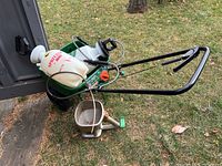 Green Scotts Turf Builder push spreader with white hopper, black metal handle, and two wheels leaning against black plastic container outdoors on grass.