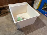 White square utility sink basin placed on cement floor, angled side view showing depth and white plastic with ribbed edges, with wrapped plumbing parts inside.