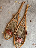 Pair of vintage wooden snowshoes laid on concrete with dry leaves scattered around.