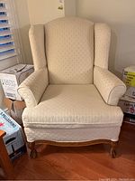 Front view of cream upholstered wingback armchair showing fabric texture, padded arms, and carved legs.