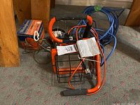 Two orange-frame work lights with black protective cages, foam handles, and multiple cords on a carpeted floor near wooden posts.