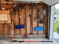 Wide view showing hanging yard tools on garage wall including two blue plastic snow shovels, various rakes, shovels, and long handled tools on wooden interior wall.