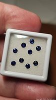 Nine round blue sapphires displayed against a white background in a small white case, showing deep royal blue color under light.