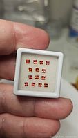 Front view showing all 15 princess cut padparadscha and papaya colored sapphires in a white display box, held by fingers for scale.