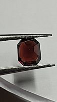 Emerald cut natural red spinel gemstone held by tweezers against a white background showing color and cut.