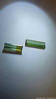 Two rectangular cut natural tourmaline gemstones, one solid green and one green to yellow bicolored, displayed under light on a plain white surface.