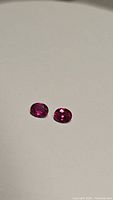 Pair of oval faceted rubies on a white/light beige background showing vivid red hot pink color and cut details.