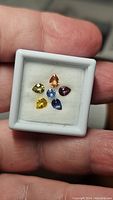 Hand-held close-up of six multicolored pear-shaped sapphires arranged in a small white display case, showcasing their colors and clarity.