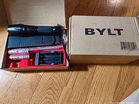 Photo of one Bylt tactical flashlight set in its original packaging with carrying case, two Ultrafire batteries, and a charger inside a cardboard BYLT box.