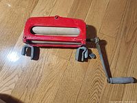 Photo of antique red metal laundry ringer showing the roller, crank handle, and clamps, placed on a wooden floor.