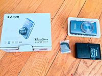 Photo showing Canon PowerShot ELPH 160 camera in blue inside protective tray, black charger, small accessory in plastic bag, and original box with some damage.