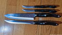 Four Cutco knives arranged on wooden floor showing blades and handles with visible use marks.