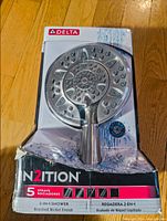 Front view of Delta In2ition 2-in-1 shower head in original worn packaging, showing circular multi-nozzle design with brushed nickel finish and product name