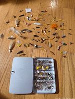 Metal fly box open with organized selection of fishing flies inside, arranged neatly by row with varying colors and feather patterns.