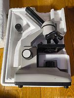LW Scientific microscope in white styrofoam packaging, showing side view including focusing knobs, stage, and eyepiece tube.