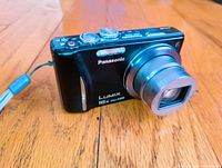 Front view of Panasonic Lumix DMC-ZS10 digital camera with extended zoom lens on wooden floor.