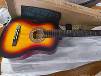 Full view of the sunburst acoustic guitar body showing the colorful sunburst finish and black bridge with white pins inside the cardboard box.
