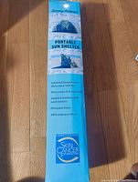 Photo of the side panel of the sealed box showing the product name Tommy Bahama Portable Sun Shelter with features including lab tested UPF 50+ sun protection, extra roomy 9-ft wide body, zippered windows with mesh interior, fiberglass frame, and 300D polyester fabric. Recommended by Skin Cancer Foundation.