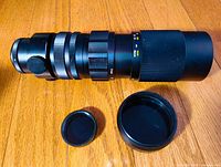 Side view of the Yashinon-R zoom lens along with two lens caps and a lens hood displayed on wooden background.