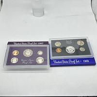 Photo of two United States proof sets from 1987 and 1968 displayed side by side in their plastic cases over respective display cards.