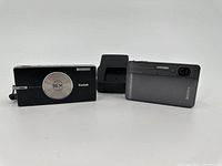 Side-by-side view of Kodak black digital camera with 5X zoom and Sony gray compact camera. Sony battery charger behind.