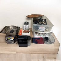 Photo showing a group of kitchen items including toaster, deep pan, rice cooker, coffee grinder, utensils bin, glass dish, and cutting boards on a wooden surface.