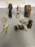 A group of eight small carvings and sculptures arranged on a flat surface showing human heads, animals, and abstract stone pieces made of stone and clay.
