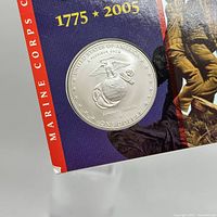 Close-up of silver-colored coin embedded in commemorative booklet featuring Marine Corps emblem with text 'United States of America Semper Fidelis One Dollar'.