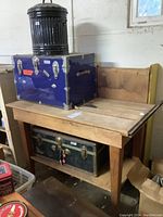Full view of wooden saw table with plank top, blue trunk on shelf, green trunk below, and black trash bin on top
