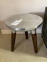 Front view of the round side table/stool showing silver-tone metal top and three tapered wooden legs.