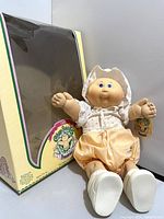 Photo of doll posed standing with original box behind it, showing box branding and doll's clothing detail.