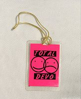 Front side of the backstage pass showing bright pink background with 'Total DEVO' text and dual smiling/frowning face graphics, glued to clear plastic with attached yellow stretchy cord.