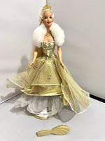 Full view of Celebration Barbie doll standing dressed in gold and white dress with gold tiara and fur stole, with brush placed beside her.