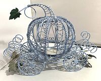 Front-left angle view of the light blue metal wire pumpkin carriage with its curved stem and leaf, showcasing the open wire pumpkin frame and large decorated wheels.