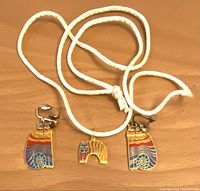 Photo of two clip earrings with colorful cat design in red, purple, blue, and orange enamel with a spiral tail, paired with cat pendant on white cord necklace.