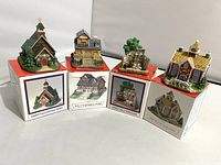 Four Liberty Falls miniature village buildings on top of their original boxes showing front and angled views. Buildings depict church, general store, mercantile and schoolhouse from a fictional 1800s mining town. Boxes show artwork of respective buildings.