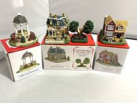 Four miniature buildings and sculptures from Liberty Falls village displayed on top of their original boxes: band stand, Kiser House, Oldest Tree, and two-story yellow roof house.