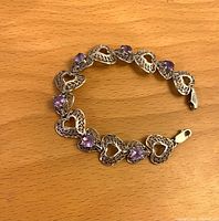 Photo of the sterling silver bracelet laid flat on a wooden surface showing alternating filigree hearts and purple heart-shaped stones.