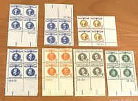 Photo showing seven blocks of four US stamps each with various colors such as blue, orange, green, and brown, showing margins with plate numbers and other markings.