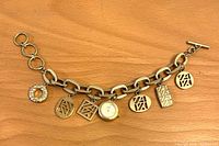 Top view of vintage Anne Klein gold tone charm bracelet with toggle clasp and integrated round watch, showing seven charms including Anne Klein logo pieces and a textured round charm.